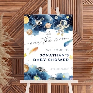 Puede incluir: Un letrero de bienvenida para baby shower sobre un caballete. El letrero presenta una ilustración de acuarela del espacio con planetas, estrellas, un astronauta y una nave espacial. El texto dice "We are over the moon. Welcome to Jonathan's Baby Shower" y la fecha.