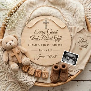Puede incluir: Un letrero de madera con el texto "Every Good And Perfect Gift COMES FROM ABOVE James 1:17 June 2028" est&aacute; rodeado por un rosario, un oso de peluche con una cruz, bloques de beb&eacute;, un body, botines de beb&eacute; y una imagen de ultrasonido.