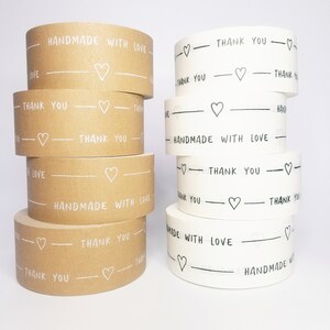 May include: Two rolls of brown and white patterned tape with the text "Handmade with Love" and "Thank You" printed on them. The tape is decorated with small hearts.