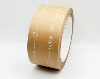 May include: A roll of brown paper tape featuring white text and heart motifs. The words "THANK YOU" are printed repeatedly along the tape. The tape is positioned at an angle against a white backdrop.