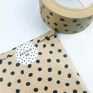 May include: A roll of brown packing tape with black polka dots. The tape is on a white surface. A brown paper bag with black polka dots and a white sticker that says "RECYCLE ME" is in the foreground.