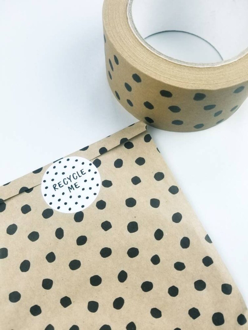 May include: A roll of brown packing tape with black polka dots. The tape is on a white surface. A brown paper bag with black polka dots and a white sticker that says "RECYCLE ME" is in the foreground.