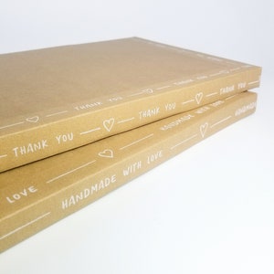 May include: Two brown cardboard boxes stacked on top of each other. The boxes are decorated with white hearts and the text "Thank You" and "Handmade with Love".