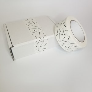 May include: A white cardboard box with a roll of white tape with a black and white geometric pattern. The tape is partially wrapped around the box.