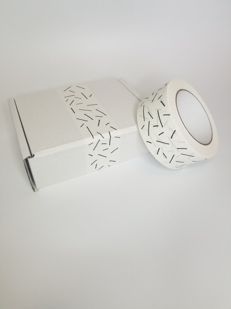 May include: A white cardboard box with a roll of white tape with a black and white geometric pattern. The tape is partially wrapped around the box.