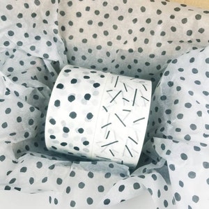 May include: Two rolls of white washi tape with black and gray geometric patterns. The tape is wrapped in tissue paper with a black polka dot pattern.
