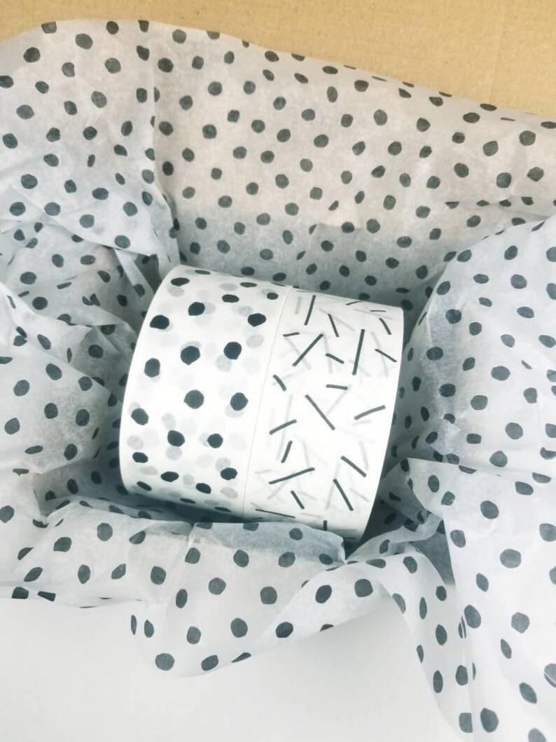 May include: Two rolls of white washi tape with black and gray geometric patterns. The tape is wrapped in tissue paper with a black polka dot pattern.