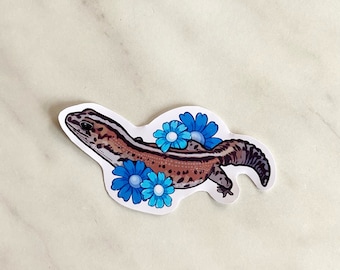 Peut inclure: Un autocollant représentant un gecko de dessin animé avec des marques brunes et blanches, entouré de fleurs bleues.