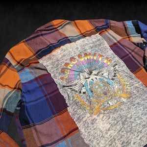 May include: A blue, orange, and purple plaid flannel shirt with a gray Aerosmith band t-shirt patch sewn on the front. The t-shirt patch features a faded graphic of a skull with wings and the text "Aerosmith" in a rainbow font.