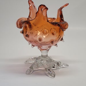 Könnte beinhalten: Eine klare Glasvase mit einer strukturierten, blasenförmigen Oberfläche. Die Vase ist hellorange und hat eine breite, offene Öffnung. Die Vase steht auf einem klaren Glasfuß mit drei Beinen.