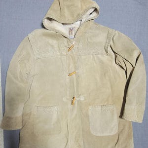 Puede incluir: Un abrigo con capucha de ante color beige con cierre de palanca y dos bolsillos delanteros. El abrigo tiene un aspecto suave y texturizado y un color cálido y neutro. La capucha está forrada con un material de color más claro.