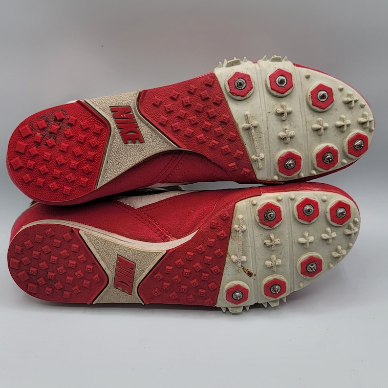 May include: A pair of red and white track shoes with metal spikes on the soles. The shoes have the Nike logo on the side.