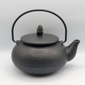 May include: Black ceramic teapot with a textured surface and a black metal handle. The teapot has a lid with a small knob on top.