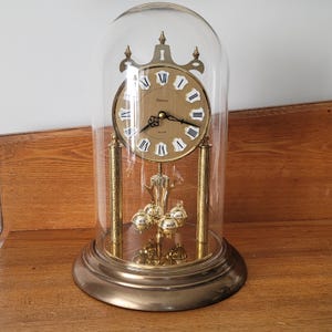 May include: A gold-coloured quartz clock encased in a glass dome. The clock face is cream with black Roman numerals and hands. The clock features a pendulum with gold spheres and sits on a round base.