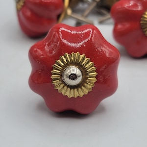 May include: A glossy red ceramic drawer knob with a scalloped, flower-like shape. The centre features a circular, metallic gold-toned decorative plate with a small, raised silver-coloured metal bolt head in the middle for attachment.