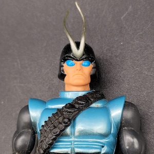 May include: A vintage action figure with a blue and black color scheme. The figure has a black helmet with clear antennae, blue eyes, and a black strap across the chest. The figure's torso is a metallic blue color.