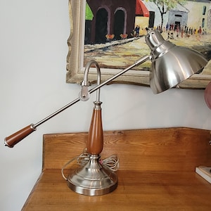 May include: A desk lamp with a silver finish, wooden accents, and an adjustable arm. The lamp features a conical shade and a wooden base. The lamp is on a wooden surface.