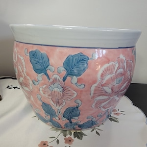 May include: A round ceramic planter featuring a white rim and a pink body decorated with blue and white floral patterns and green leaves. The exterior surface displays a repeating motif of stylised blossoms and foliage in a soft colour palette.