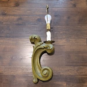 May include: A wall-mounted sconce lamp featuring an olive green, C-shaped scrollwork base with carved leaf-like details. The fixture includes a white, spiraled candle-style stem, a brass-toned socket, and a single white LED light bulb.