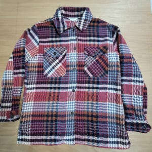 May include: A long-sleeved button-down shirt with a plaid pattern in shades of red, white, black, and blue. The shirt has two chest pockets and a collared neckline. The cuffs have buttons.