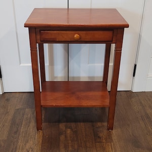 Peut inclure: Une table d'appoint rectangulaire en bois avec une finition marron chaud. La table présente une surface supérieure plate, un tiroir horizontal unique avec un petit bouton rond en bois et une étagère inférieure ouverte sur quatre pieds droits.
