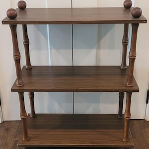 May include: A three-tier wooden shelving unit with a dark brown wood grain finish. The structure features four vertical supports with a turned, bamboo-like design and four decorative wooden spheres positioned at the top corners.