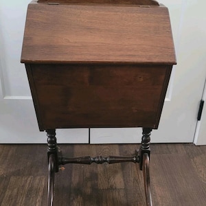 May include: A dark brown wooden sewing box with a hinged lid and a handle. The box is mounted on a stand with decorative legs. The stand has a horizontal bar connecting the legs. The box is approximately 76 cm tall.