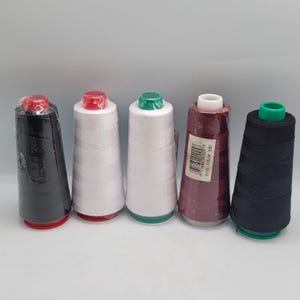 Peut inclure: Cinq bobines coniques de fil à coudre disposées en rangée. Les couleurs incluent noir avec un capuchon rouge, blanc avec un capuchon rouge, blanc avec un capuchon vert, bordeaux foncé avec un capuchon blanc et noir avec un capuchon vert.