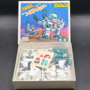 Puede incluir: Una caja de kit de modelo vintage con una ilustración de robot y el texto "VF-1S". La caja es blanca con detalles azules y rojos. Dentro de la caja abierta hay piezas de modelo de plástico sin montar y pegatinas.