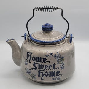 Peut inclure: Une théière en céramique beige mouchetée avec un couvercle et un bord bleus. L'avant présente le texte "Home Sweet Home" en lettres bleues, entouré d'illustrations bleues de papillons et de fleurs. Elle possède une anse en métal noir.