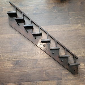 Peut inclure: Une étagère murale en forme d'escalier en bois teinté foncé avec sept marches et une rampe. L'étagère comporte six petits crochets noirs pour suspendre des objets. Le grain du bois est visible.