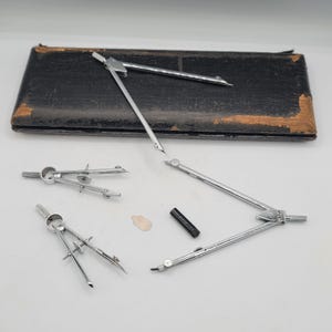Könnte beinhalten: Eine Sammlung silberner Zeichenzirkel und Werkzeuge, darunter ein großer Zirkel, kleinere Zirkel, ein schwarzes zylindrisches Teil und ein kleines, beiges, flaches Teil. Die Werkzeuge sind auf einer weißen Oberfläche mit einem schwarzen Lederetui im Hintergrund angeordnet.