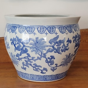 May include: A large, round ceramic planter with a white base and a wide, slightly flared rim. The planter is decorated with blue floral and foliate patterns. The rim features a repeating heart-shaped design. The planter sits on a wooden surface.