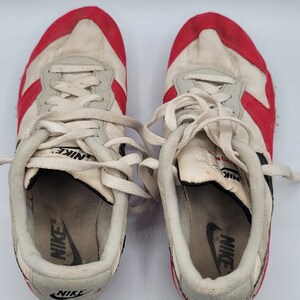 May include: A pair of white and red Nike running shoes with white laces. The shoes have a black Nike logo on the side and the word "Nike" printed on the tongue.