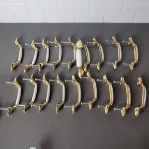 May include: A collection of 18 gold-toned cabinet knobs with white ceramic inserts. The knobs have a simple, classic design and are arranged in rows on a white tiled surface.