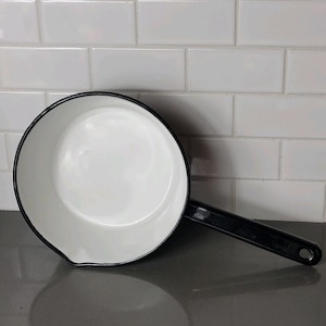 Peut inclure: Une casserole en émail blanc avec un bord et une poignée noirs. La casserole a un bec verseur et un trou au bout de la poignée pour l'accrocher. L'intérieur est blanc et l'extérieur est noir.