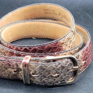 May include: A genuine imported python belt with a gold-coloured buckle. The belt features a snakeskin pattern in shades of brown, tan, and burgundy. The interior of the belt is a light tan colour, and the text "Genuine Imported Python" is visible.