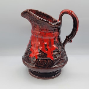 May include: A vintage ceramic pitcher with a dark brown base and handle, and a vibrant red and brown lava glaze. The pitcher has a rounded body, a spout, and a curved handle. It is a decorative piece.