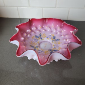 Può includere: Una ciotola di vetro rosa e bianca con un bordo a festoni e un motivo floreale. La ciotola ha una superficie testurizzata ed è decorata con piccoli punti bianchi.