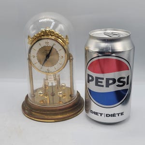 May include: A gold-toned anniversary clock with a white face, black hands, and a rotating pendulum, enclosed in a glass dome on a circular wooden base, positioned next to a silver 355ml Diet Pepsi aluminium soda can for scale reference.