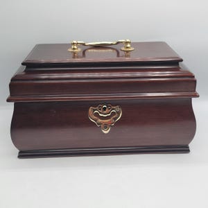 May include: A dark brown wooden box with a polished finish, featuring a gold-toned metal handle on the top lid and a decorative gold-toned metal keyhole plate centered on the front panel. The box has a rectangular shape with tiered edges.