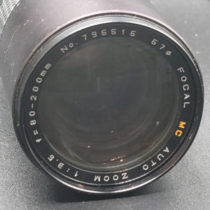 Könnte beinhalten: Ein schwarzes Zoomobjektiv mit der Aufschrift "Focal MC Auto Zoom" und "f=80-200mm 1:3.5". Das Objektiv hat einen Durchmesser von 67 mm und die Seriennummer 796516. Das Objektiv ist für die Fotografie konzipiert.