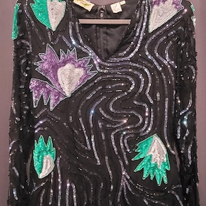 Op de afbeelding: Zwarte top met pailletten en een V-hals, versierd met zilveren, groene en paarse bloemmotieven. De blouse met lange mouwen is versierd met pailletten en hangt aan een grijze hanger.