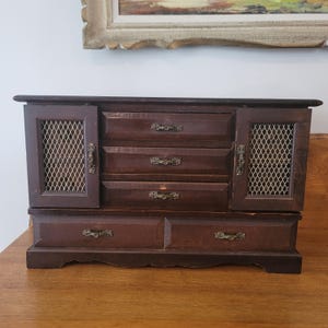 Puede incluir: Un joyero de madera de color marrón oscuro con tres cajones centrales apilados y dos armarios laterales con paneles de malla metálica. Cada cajón y puerta de armario tiene pequeños tiradores de metal de estilo antiguo en tono latón.
