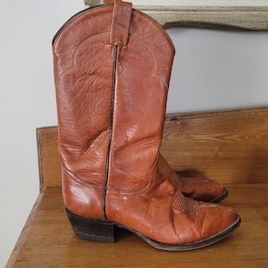 May include: A pair of brown leather cowboy boots. The boots feature a classic design with a pointed toe, decorative stitching, and a stacked heel. The leather has a worn appearance, suggesting they are vintage.
