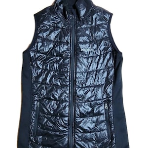 Peut inclure: Gilet matelassé noir Marmot avec fermeture éclair et deux poches zippées. Le gilet a un col montant et une doublure en polaire noire.