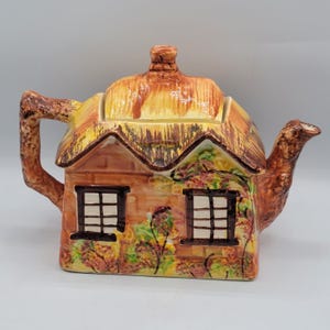 Peut inclure: Théière en céramique en forme de cottage. Elle présente un toit de chaume, des murs bruns avec deux fenêtres, un bec verseur et une anse. Les couleurs sont le marron, le jaune et le vert, pour un aspect rustique. Idéale pour préparer le thé.