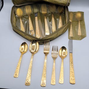 Könnte beinhalten: Ein goldfarbenes Besteckset mit einem floralen Prägemuster auf den Griffen. Die Kollektion umfasst verschiedene Löffel, Gabeln und ein Messer, wobei einige Teile in einer gesteppten, olivgrünen Reißverschlusstasche mit Klarsichthüllen aufbewahrt sind.