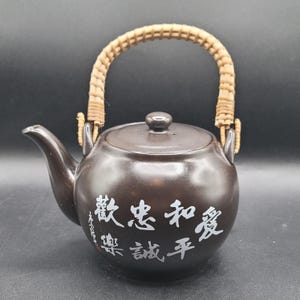 May include: A brown ceramic teapot with a woven handle and a lid. The teapot has white Japanese characters on the front. The spout is curved, and the handle is made of woven light brown material.
