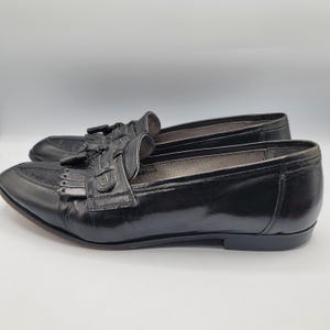 May include: Pair of black leather loafers with a glossy finish. The shoes feature a decorative tassel and fringe detail on the vamp. The soles are a dark brown color.